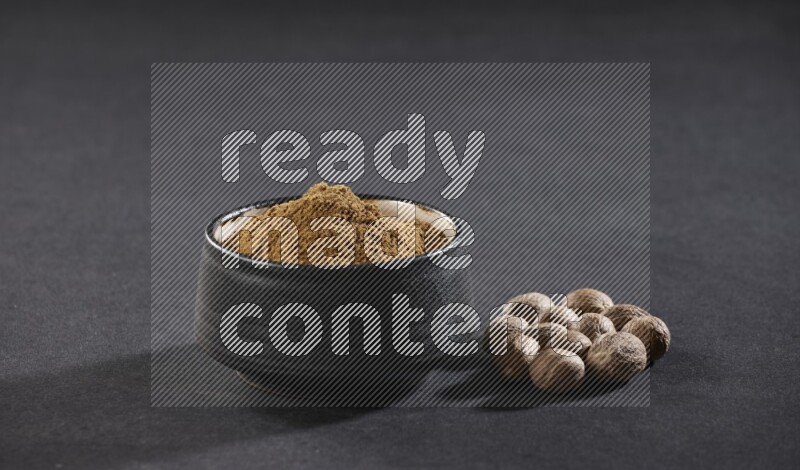 وعاء فخاري أسود ممتلئ ببودرة جوزة الطيب مع بعض حبوب جوزة الطيب بجانبه على خلفية سوداء