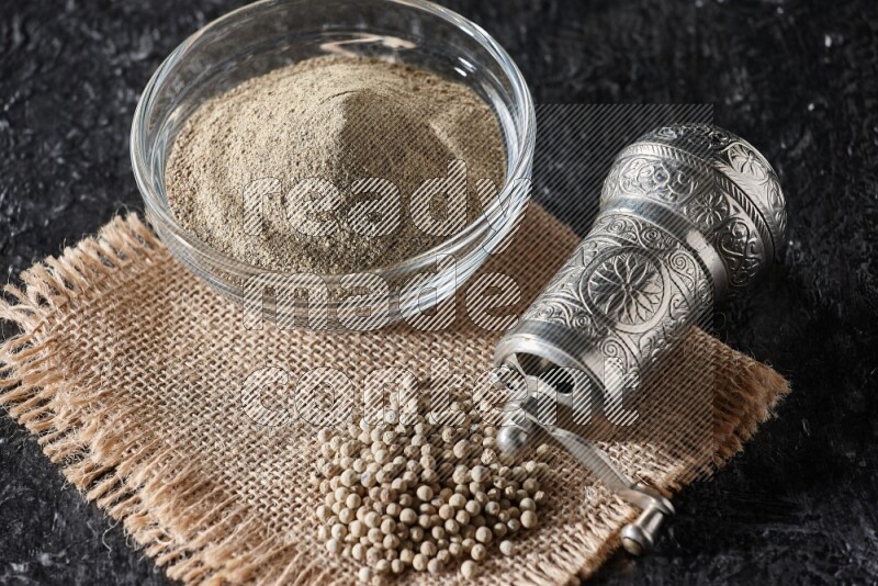A glass bowl full of white pepper powder with white pepper beads on a burlap piece of fabric and a metal grinder on textured black flooring