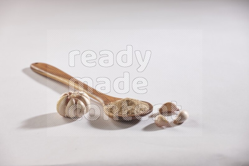 A wooden ladle full of garlic powder with a whole garlic bulb and some cloves beside it on a white flooring