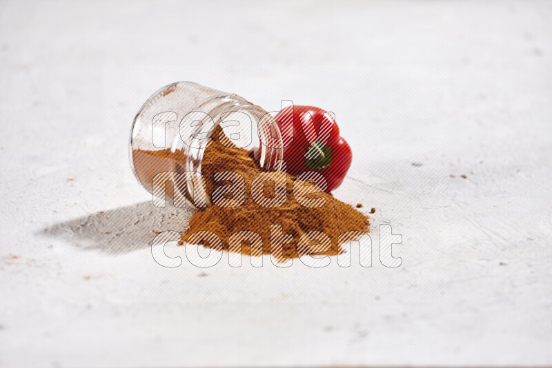 A glass jar full of ground paprika powder flipped with some spilling powder on white background