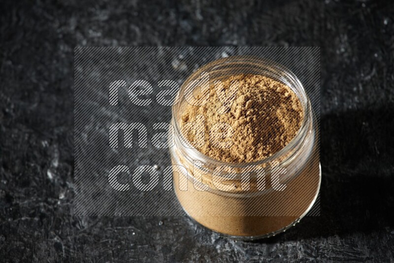 A glass jar full of allspice powder on a textured black flooring