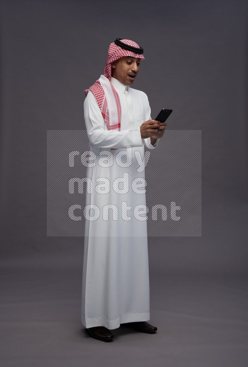 Saudi man wearing thob and shomag standing texting on phone on gray background