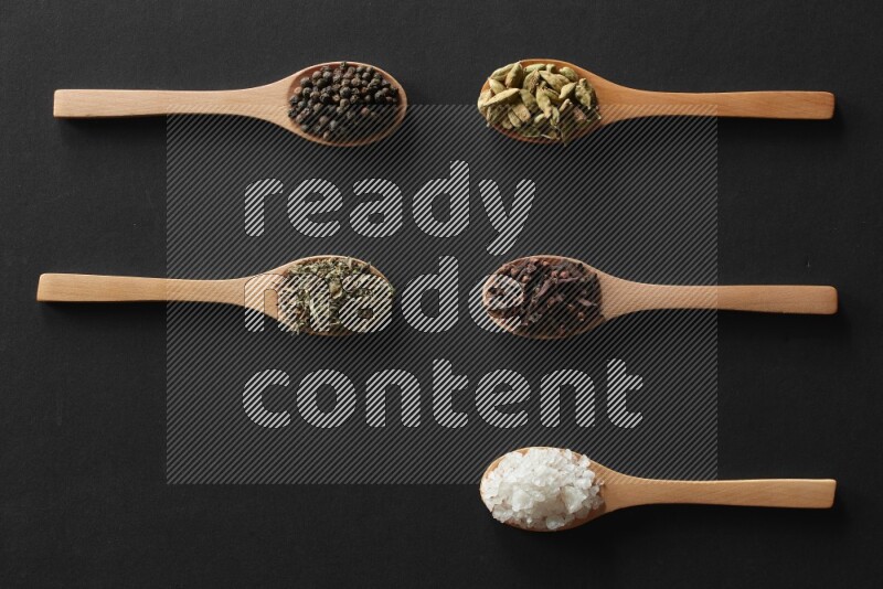 wooden spoons filled with white peppers, cloves, cardamom, salt, black peppers and basil on black flooring