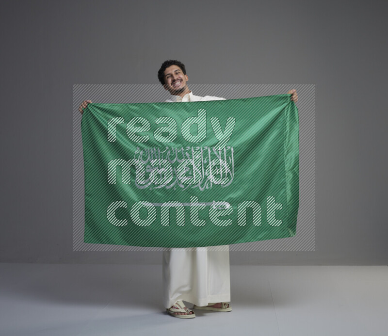 A Saudi man standing wearing thob holding big Saudi flag on gray background