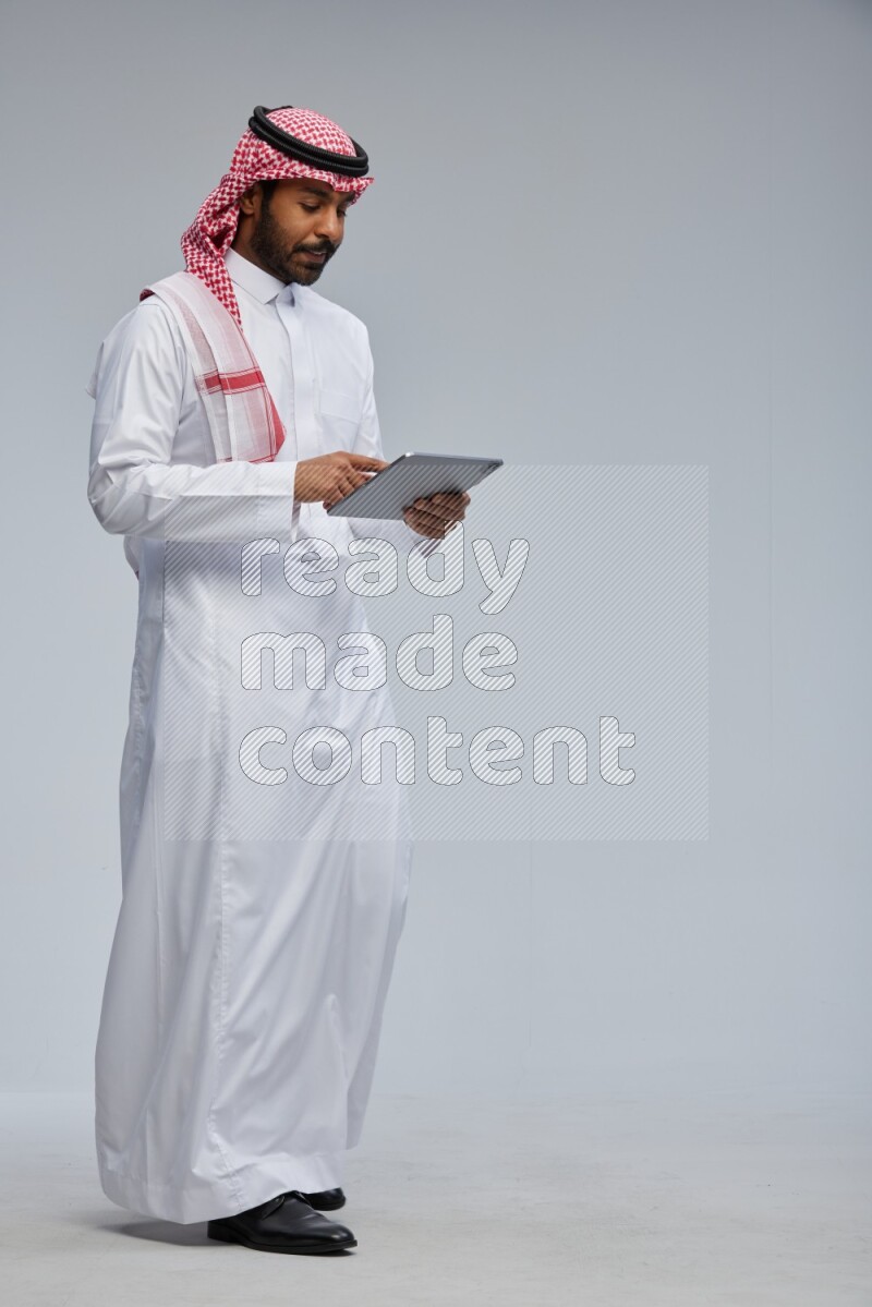 Saudi man Wearing Thob and shomag standing working on tablet on Gray background