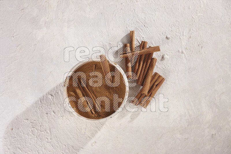Ceramic bowl full of cinnamon powder with cinnamon sticks on the side on white background