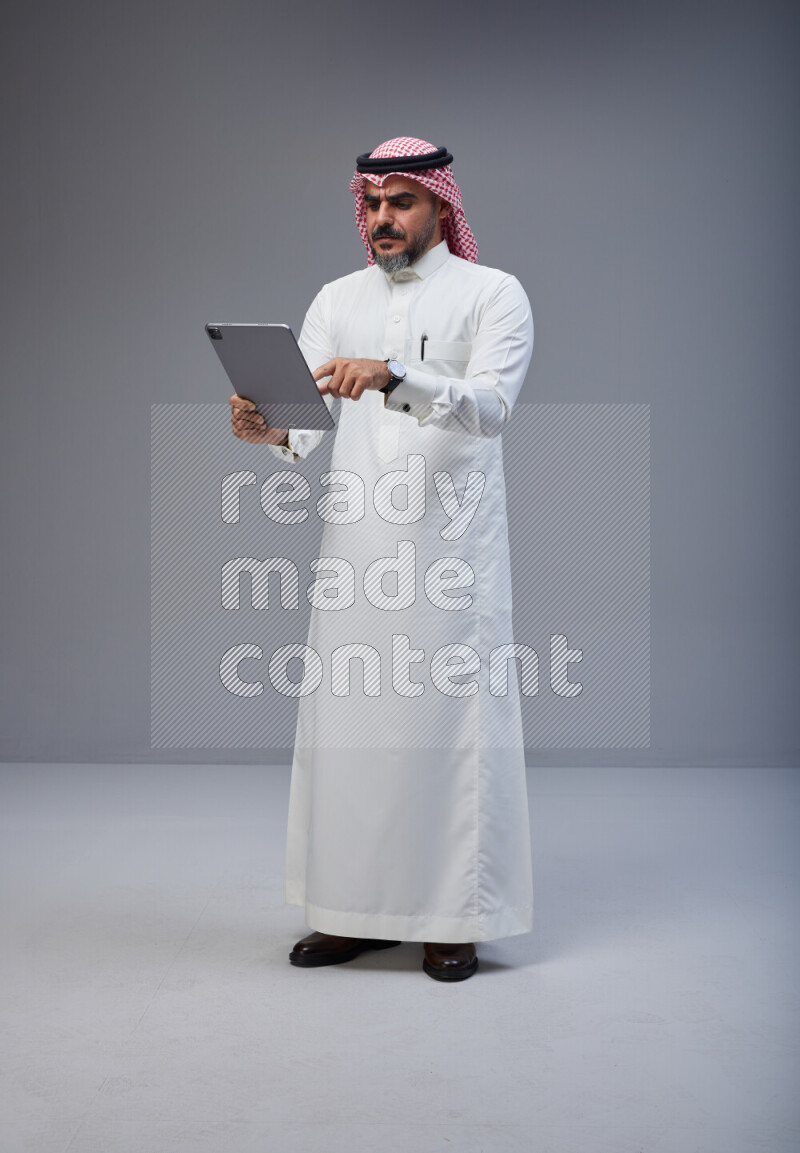 Saudi man Wearing Thob and red Shomag standing working on tablet on Gray background