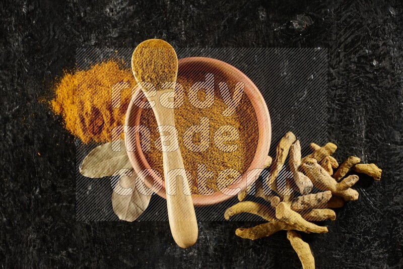 A wooden bowl and a wooden spoon full of turmeric powder with dried turmeric fingers on textured black flooring