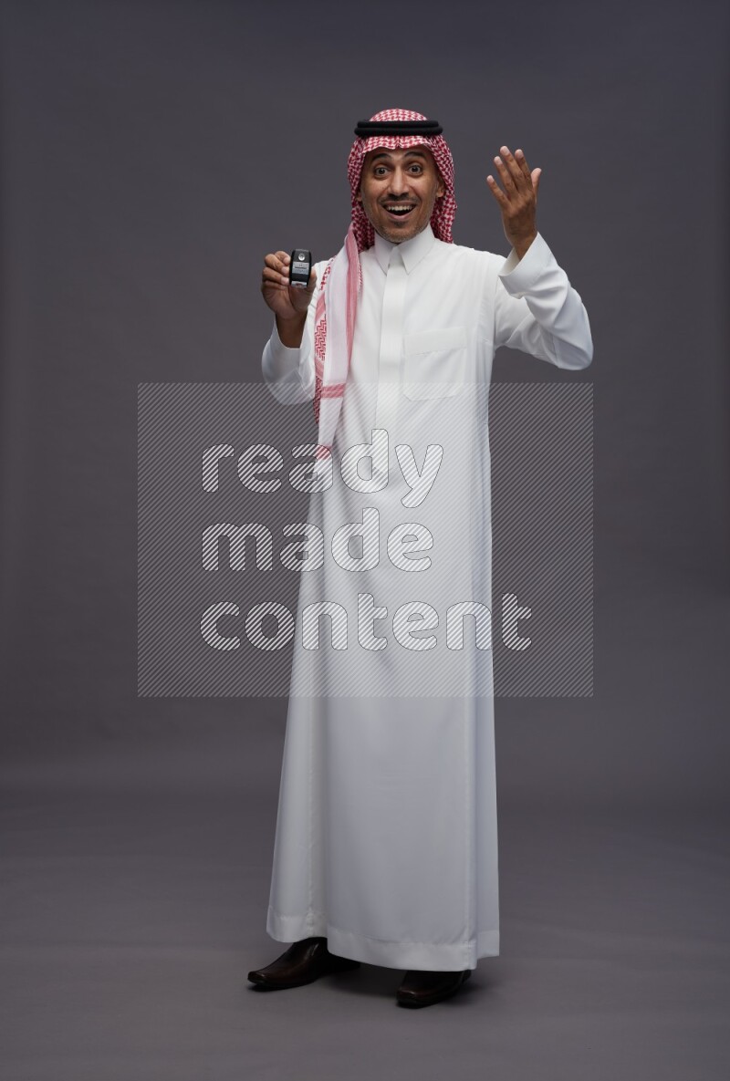 Saudi man wearing thob and shomag standing holding key car on gray background