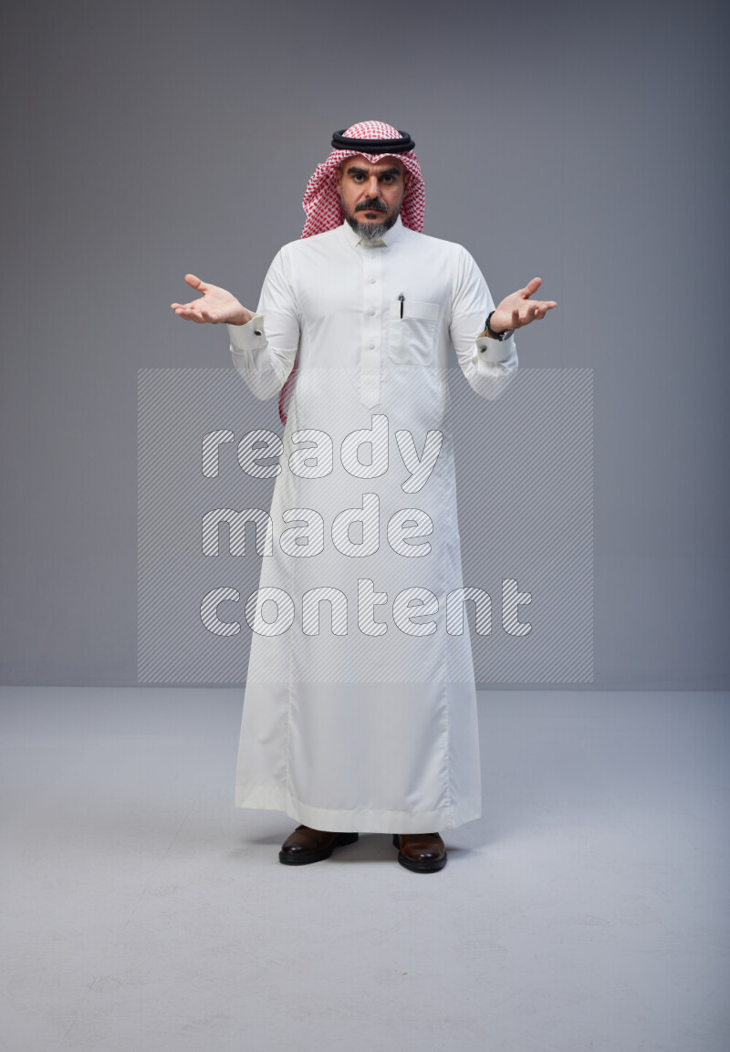 Saudi man Wearing Thob and red white Shomag standing interacting with the camera on Gray background
