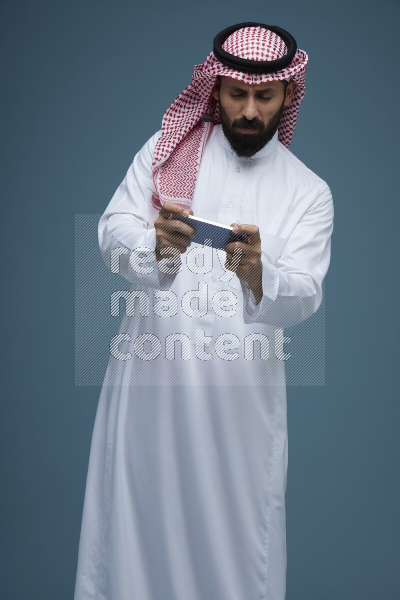 A man Playing a Game on his phone in a blue background wearing Saudi Thob with Shomag