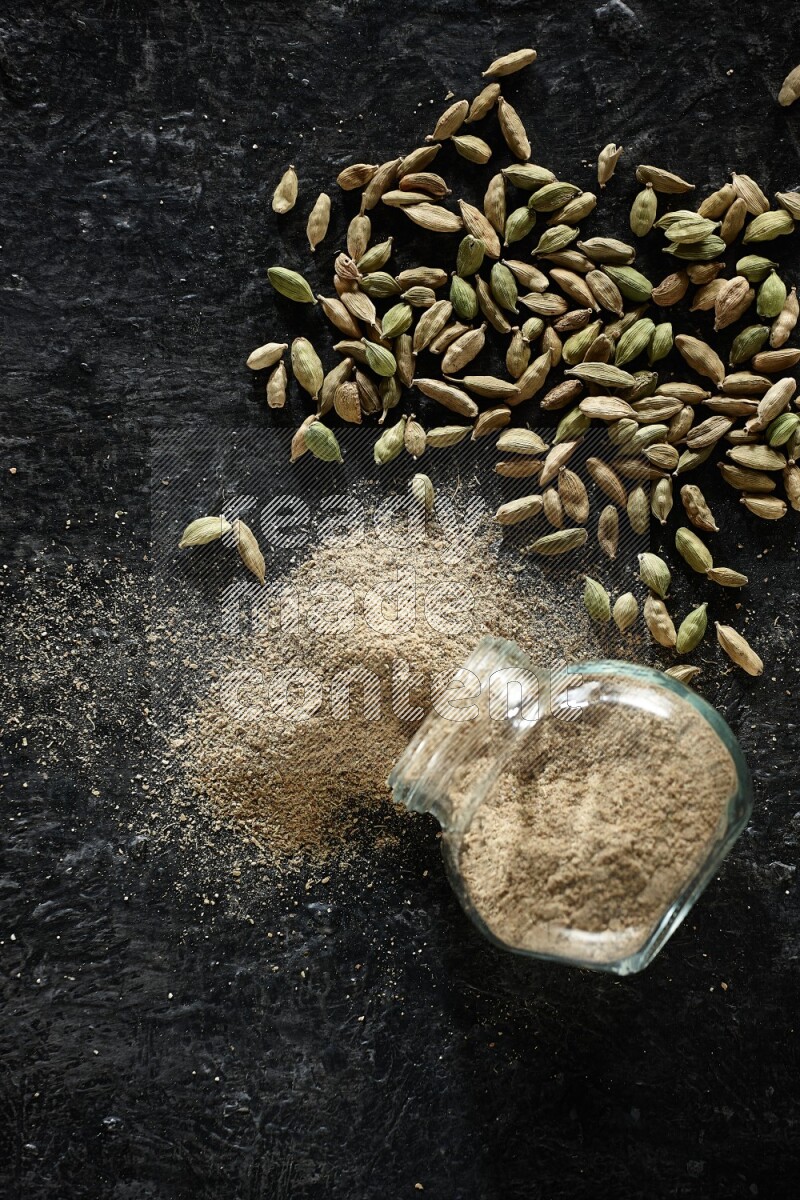 A flipped glass spice jar full of cardamom powder and powder spilled out of it with cardamom seeds spreaded on textured black flooring