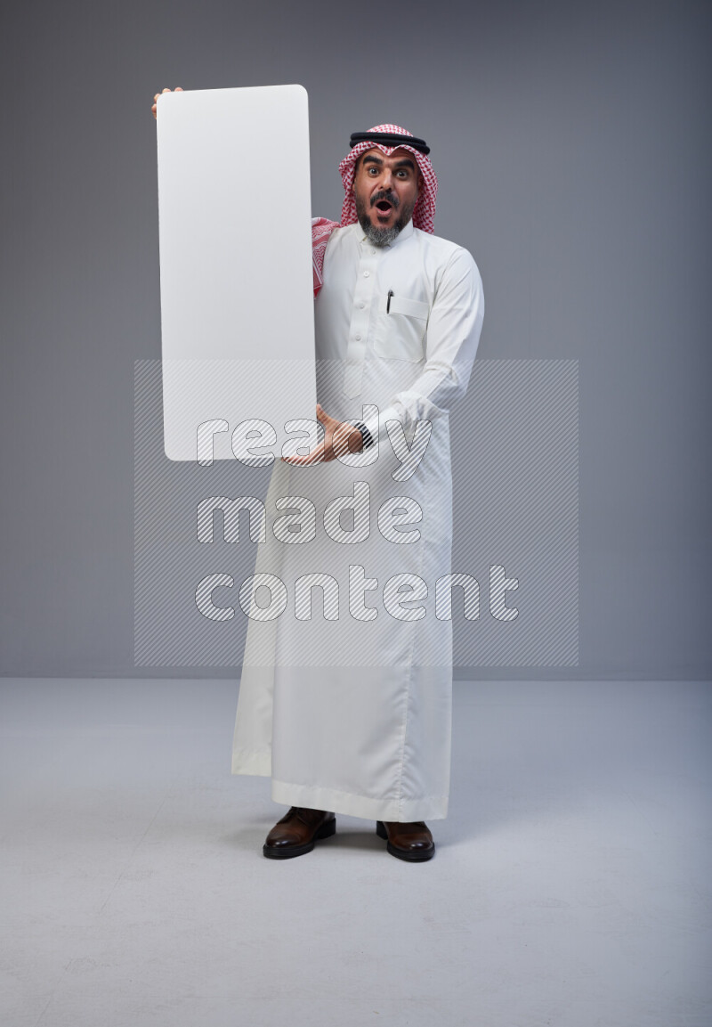 Saudi man Wearing Thob and red Shomag standing holding board on Gray background