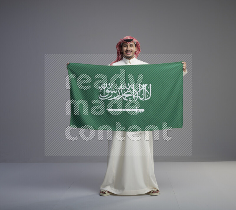 A saudi man standing wearing thob and red shomag holding big saudi flag on gray background