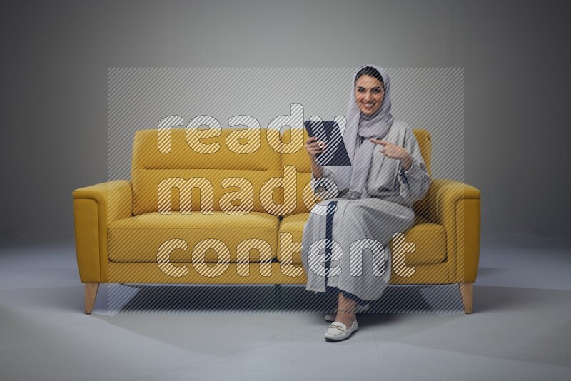 A Saudi woman wearing a light gray Abaya and head scarf sitting on a yellow sofa and holding her tablet while pointing to it eye level on a grey background