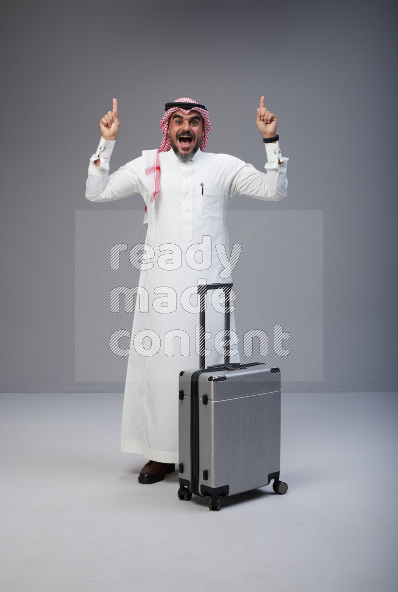 Saudi man wearing Thob and red Shomag standing holding Travel bag on Gray background