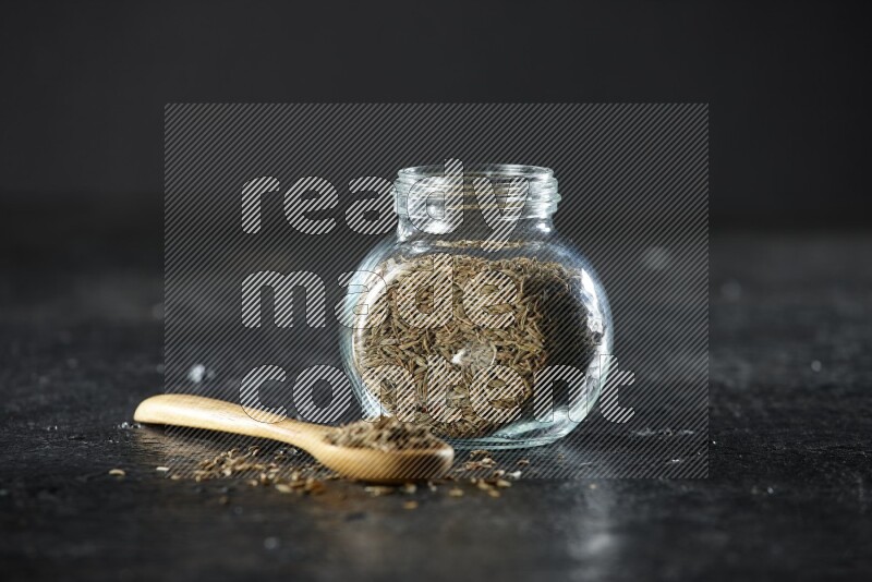 A glass spice jar and a wooden spoon full of cumin seeds on a textured black flooring