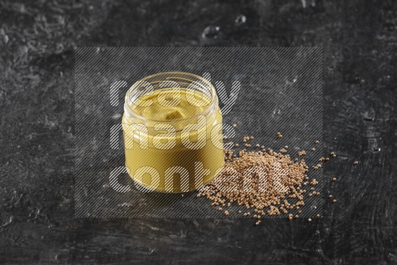 A glass jar full of mustard paste and mustard seeds spread next to it on a textured black flooring