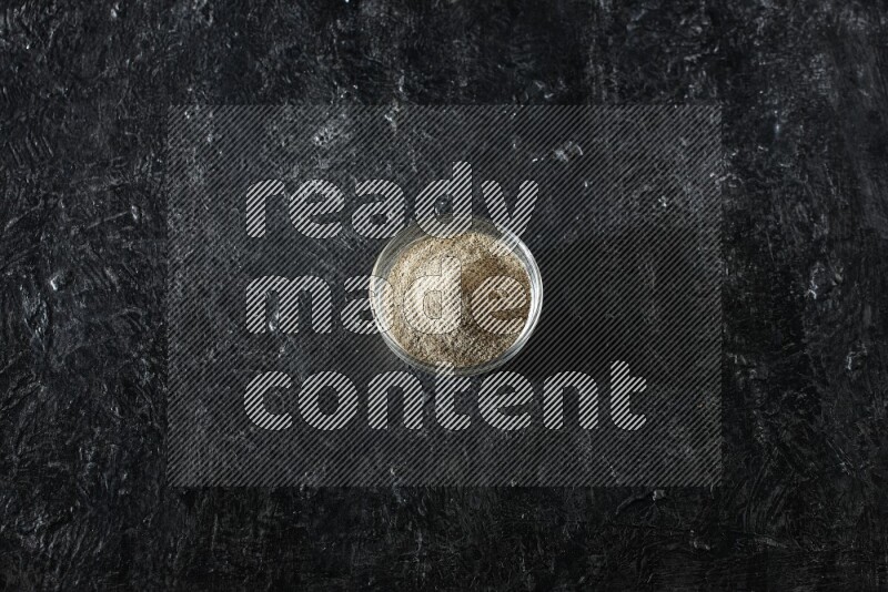 A glass bowl full of cardamom powder on textured black flooring