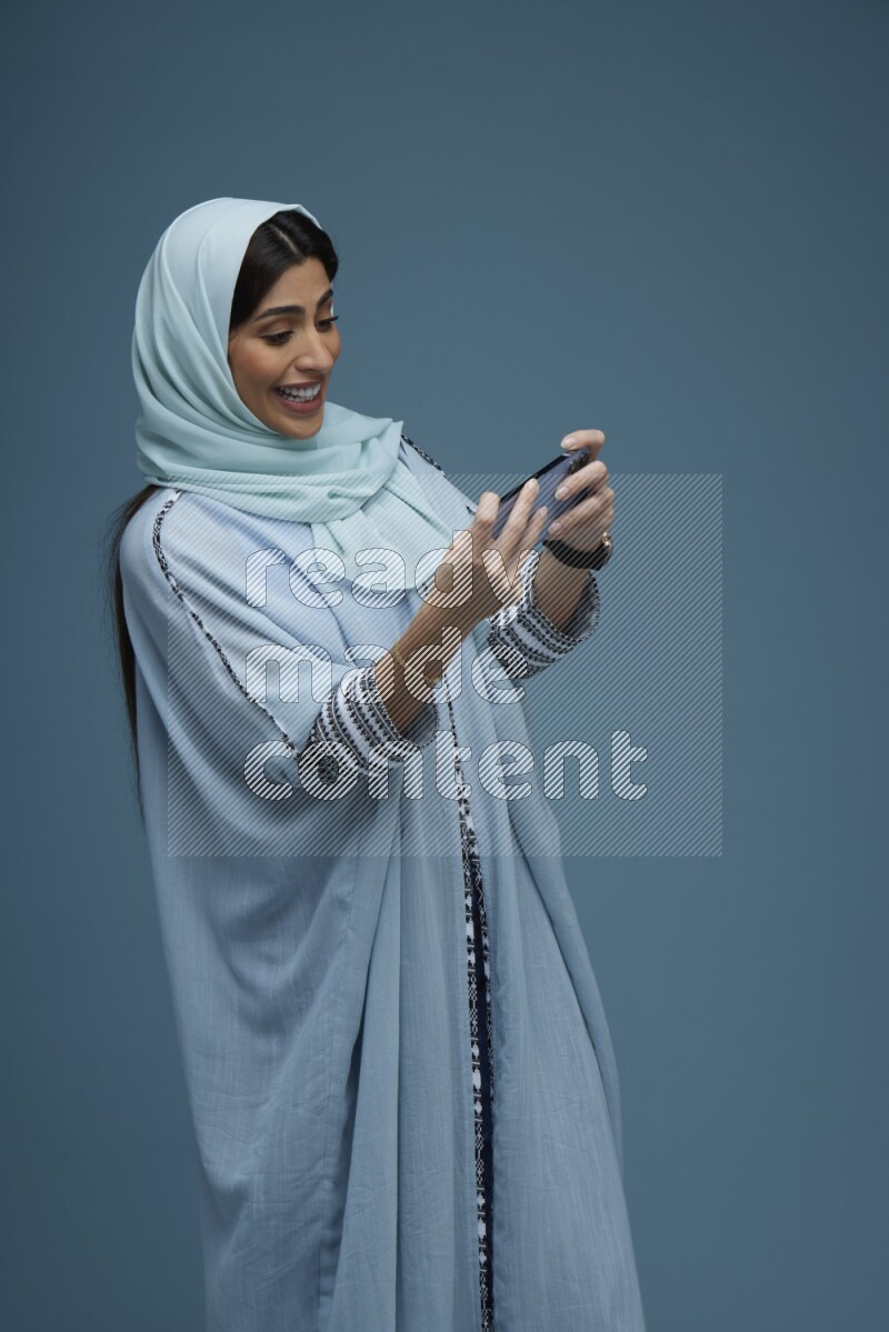 A Saudi woman Playing a Game on her phone on a blue background wearing a blue Abaya with hijab