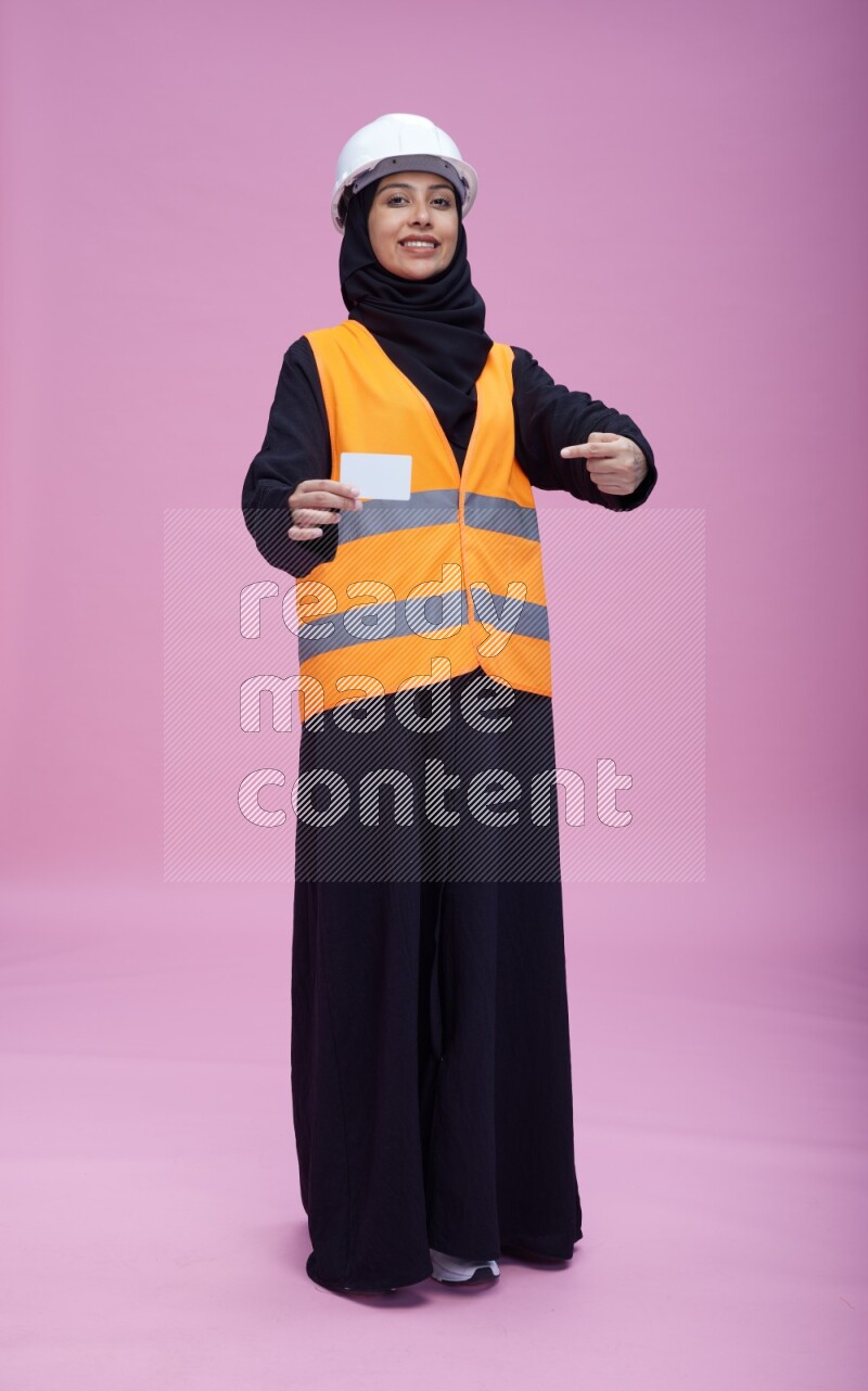 Saudi woman wearing Abaya with engineer vest and helmet standing holding ATM card on pink background