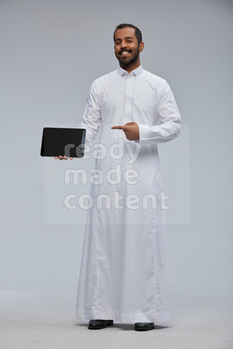 Saudi man wearing Thob standing showing tablet to camera on Gray background
