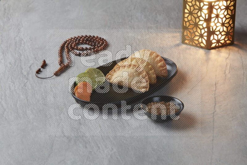 Three fried sambosas in an oval shaped black plate and a red sauce in a black round ramekin with a brown misbaha and a golden lantern on the side on a gray background