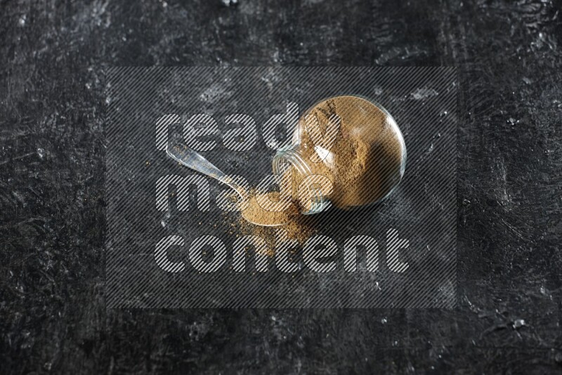 A flipped glass spice jar and a metal spoon full of cumin powder and powder spilled out on a textured black flooring