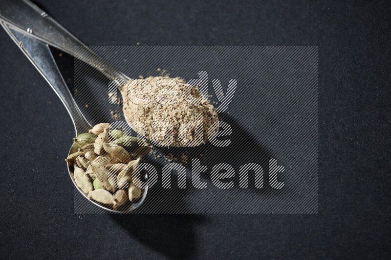 ملعقتان معدنيتان ممتلئتان ببودرة الهيل وحبوب الهيل على خلفية سوداء