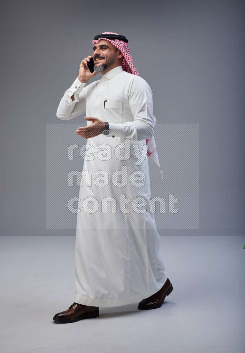 Saudi man Wearing Thob and red Shomag standing talking on phone on Gray background