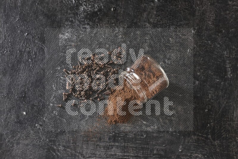 وعاء زجاجي مقلوب ممتلئ ببودرة القرنفل مع حبوب القرنفل المتناثرة على أرضية سوداء