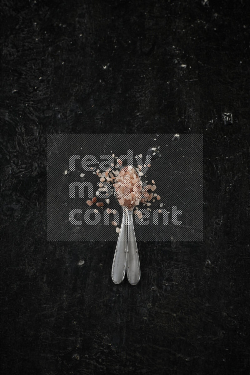 2 metal spoons filled with pink himalayan salt on black background