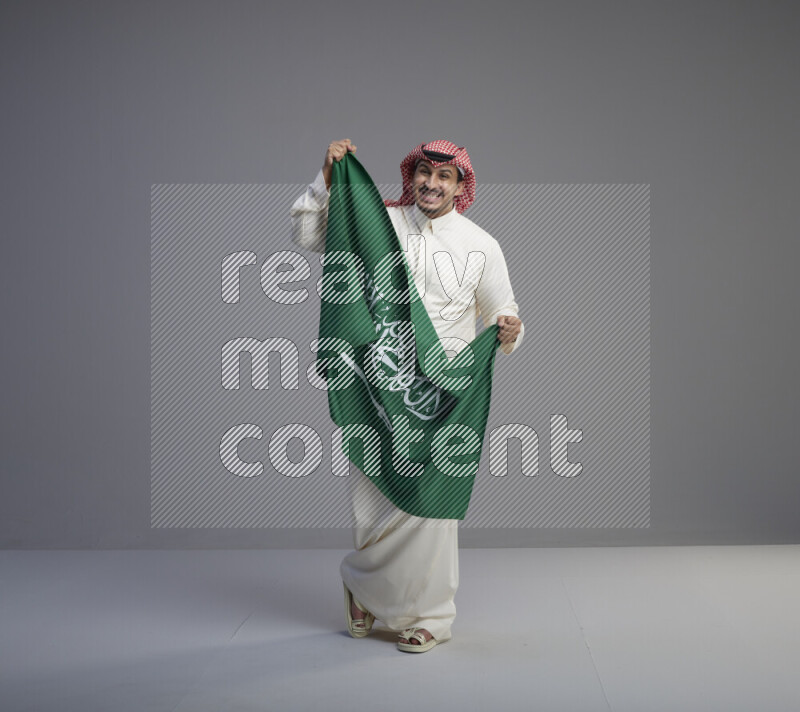 A saudi man standing wearing thob and red shomag holding big saudi flag on gray background