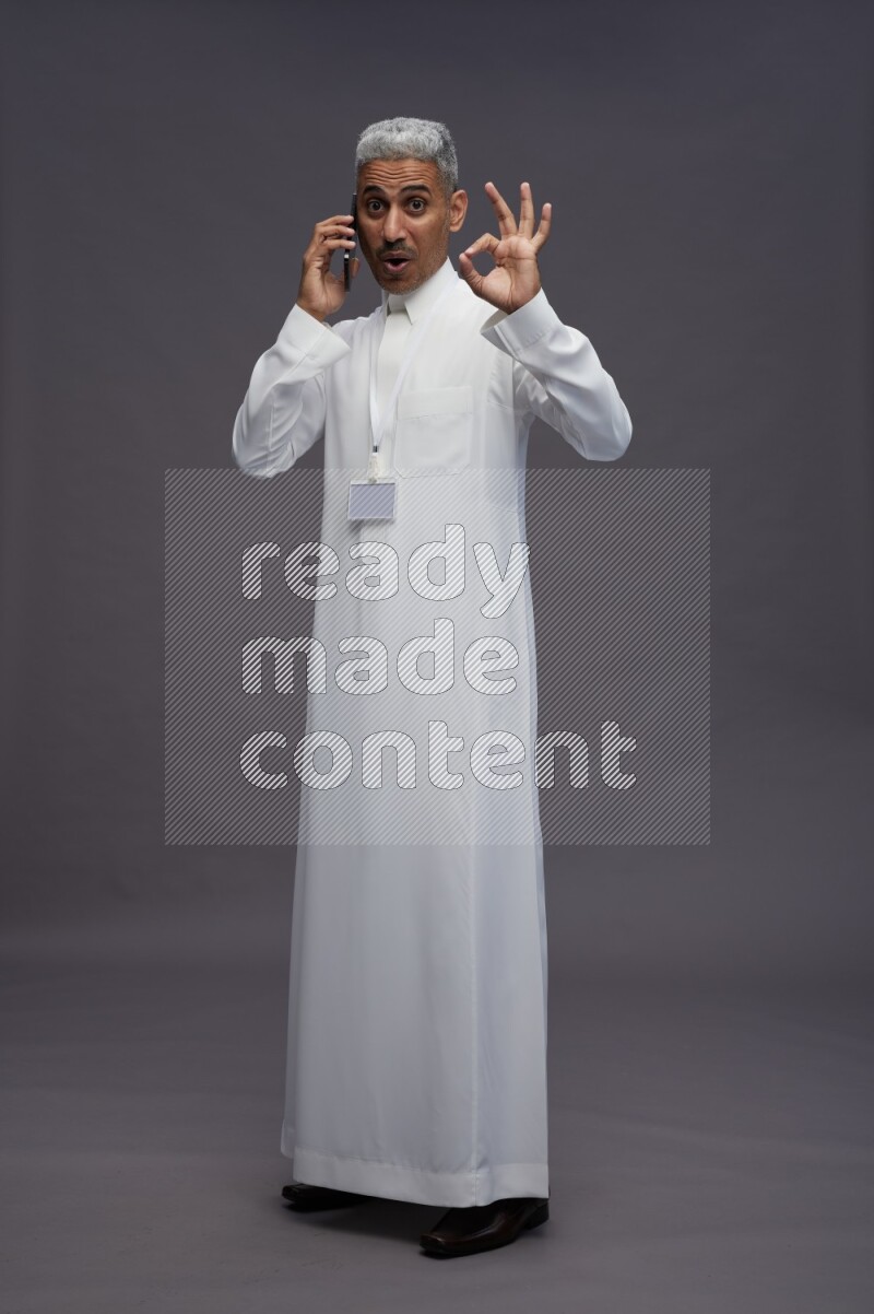 Saudi man wearing thob with neck strap employee badge standing talking on phone on gray background