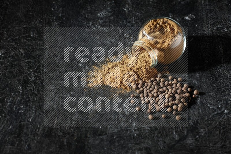 وعاء زجاجي للتوابل مقلوب ممتلئ ببودرة بهار الفلفل الحلو والبودرة تناثرت منه مع الحبوب على خلفية سوداء