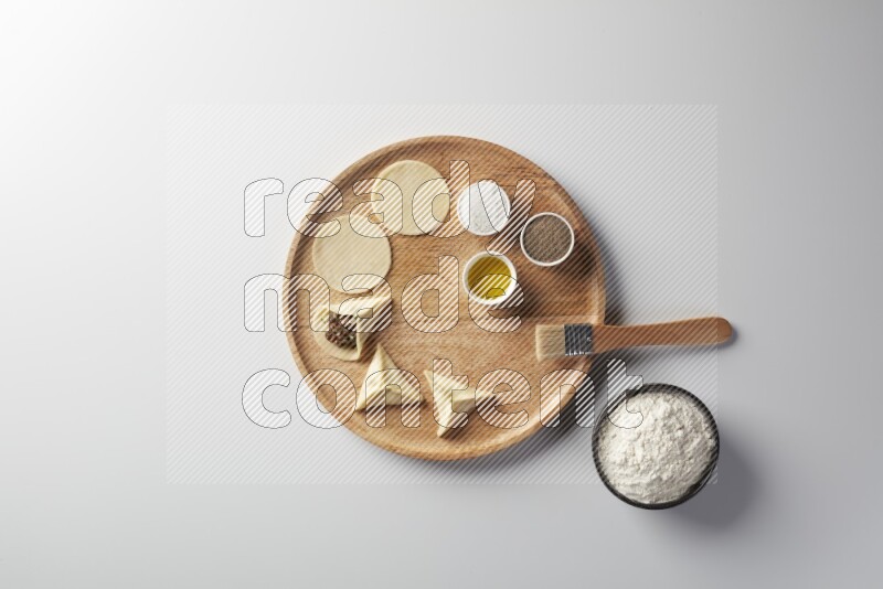 two closed sambosas and one open sambosa filled with meat while flour, salt, black pepper and oil with oil brush aside in a wooden dish on a white background