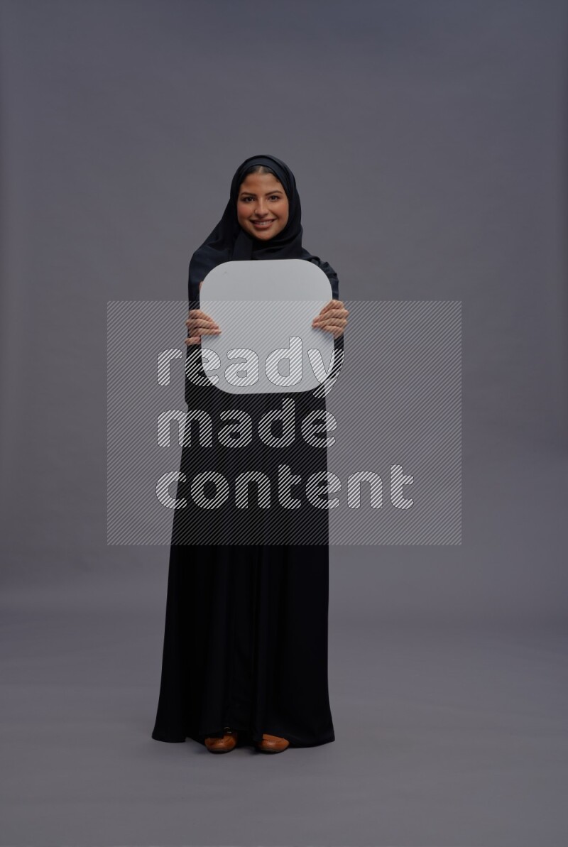 Saudi woman wearing Abaya standing holding social media sign on gray background