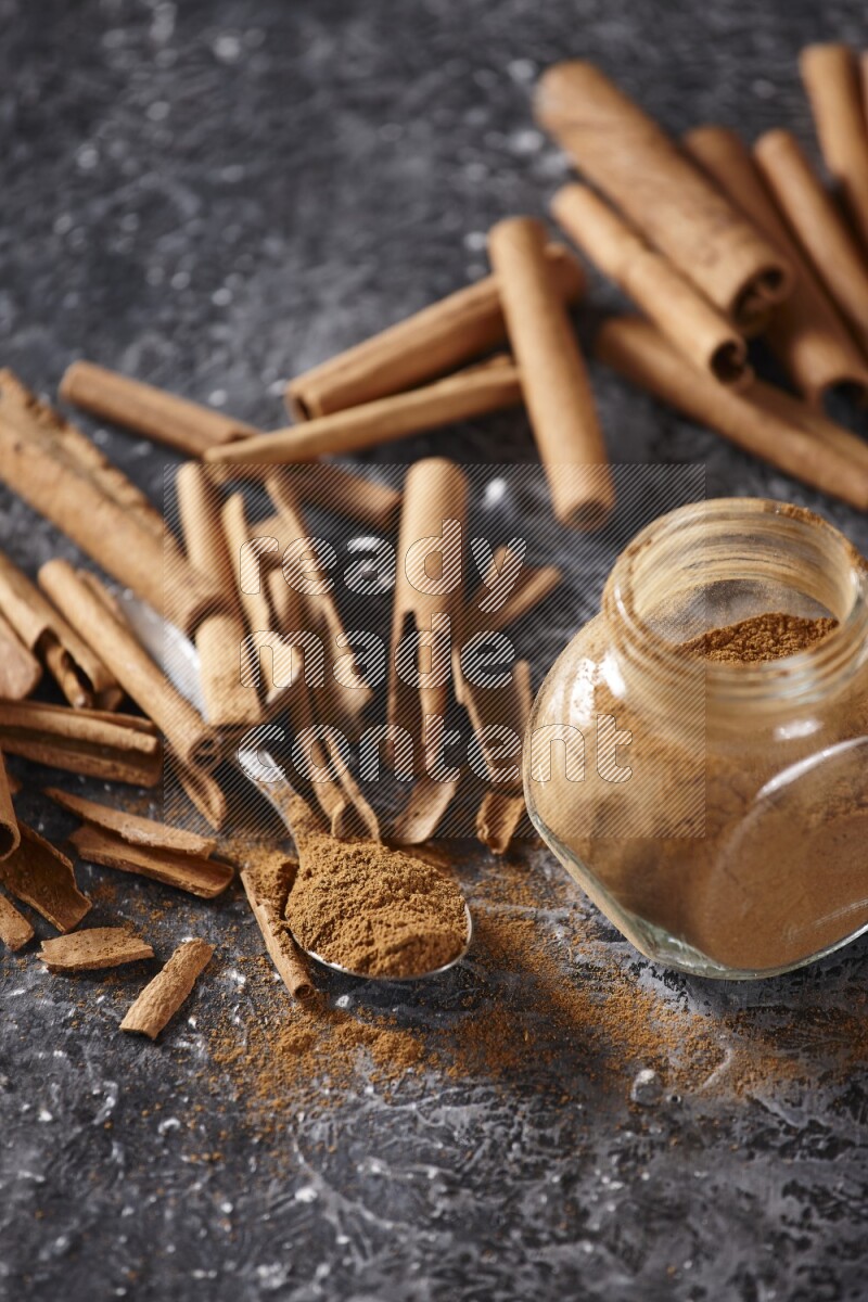 Herbal glass jar and a metal spoon full of cinnamon powder surrounded by cinnamon sticks on textured black background