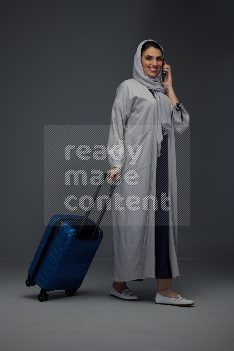 A Saudi woman wearing a light gray Abaya and head scarf standing and talking in the phone while holding shopping bags eye level on a grey background