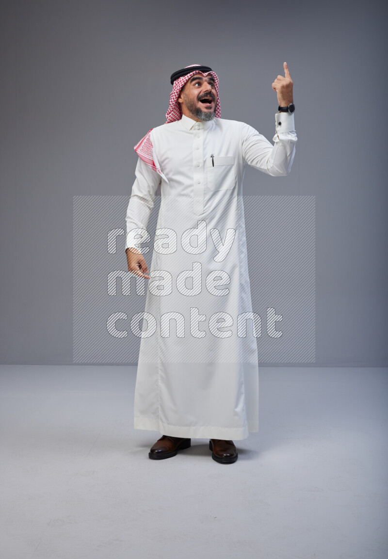 Saudi man Wearing Thob and red white Shomag standing interacting with the camera on Gray background