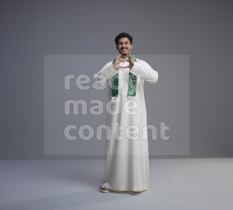 A Saudi man standing wearing thob and Saudi flag scarf on gray background