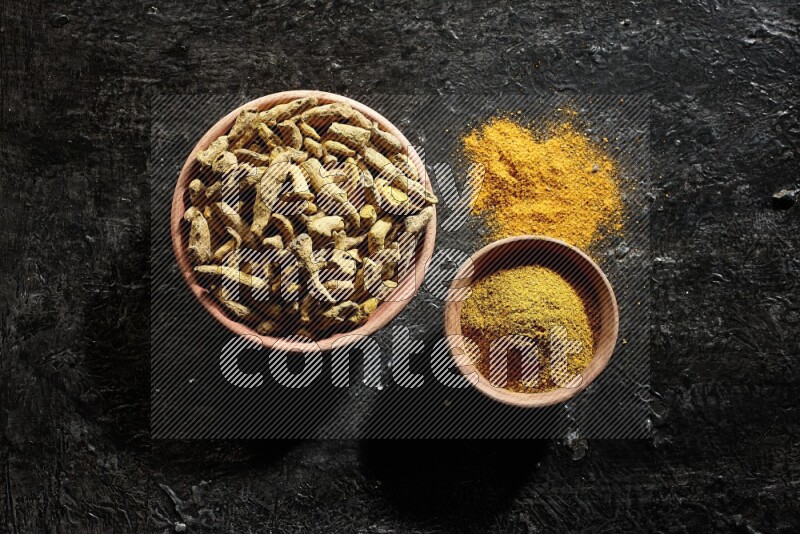 2 wooden bowls, one full of turmeric powder and the other full of dried turmeric whole fingers on a textured black flooring