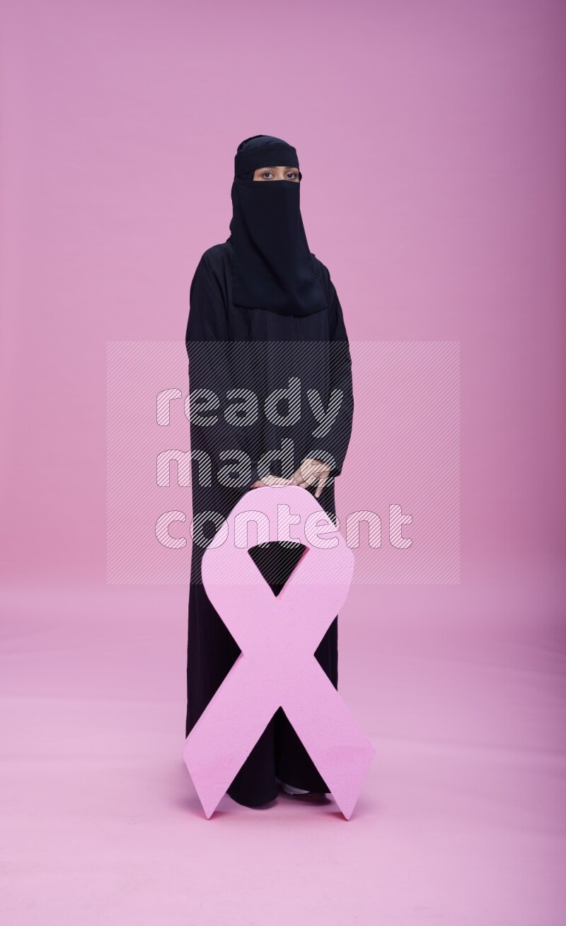 Saudi woman wearing abaya and niqab standing holding awareness ribbon on pink background