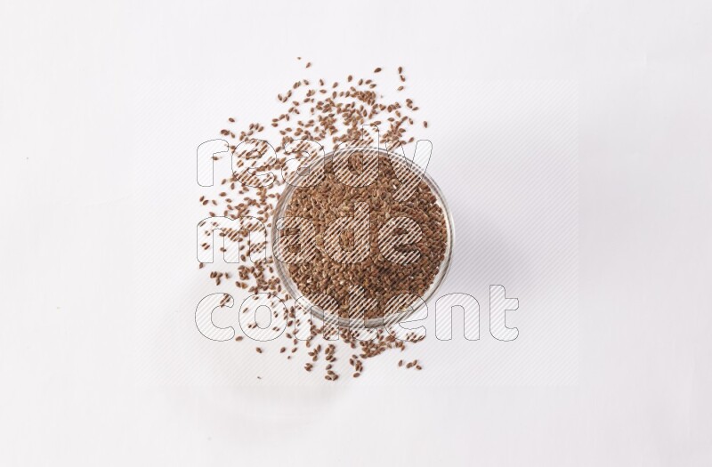 A glass bowl full of flax seeds surrounded by flax seeds on a white flooring