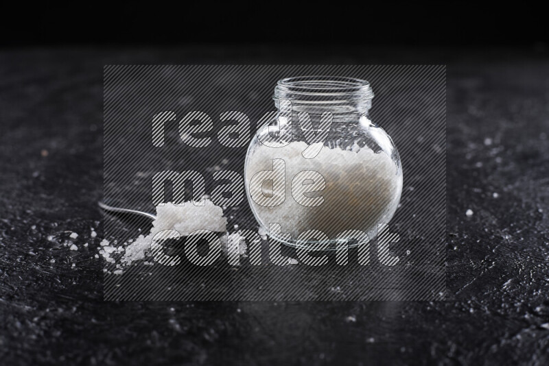 A glass jar full of coarse sea salt crystals on black background