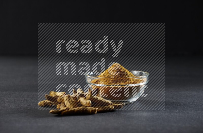 A glass bowl full of turmeric powder and dried whole fingers beside it on black flooring
