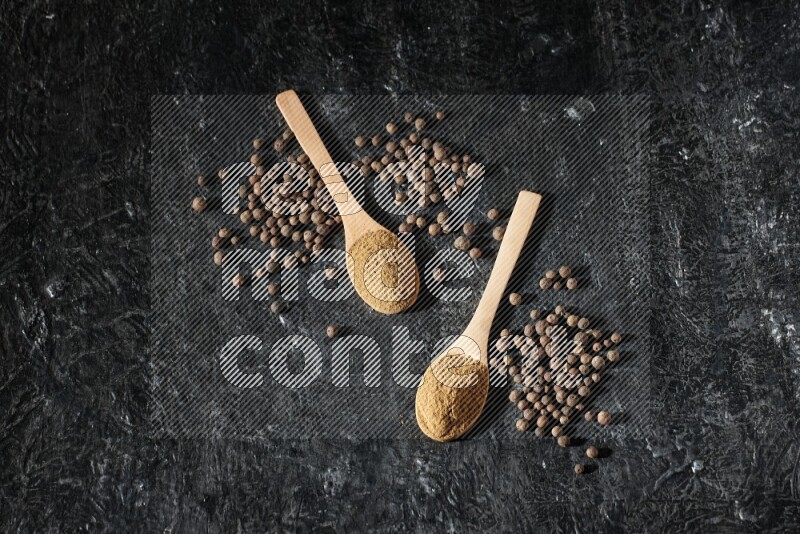 Wooden spoons full of all spice powder and allspice whole balls beside it on a textured black flooring