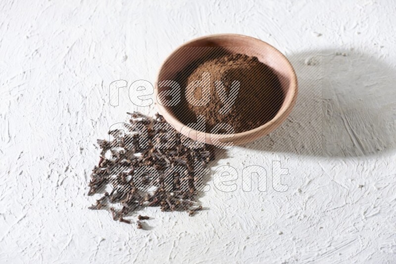 A wooden bowl full of cloves powder with whole cloves beside it on a textured white flooring