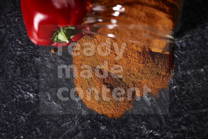 A glass jar full of ground paprika powder flipped with some spilling powder on black background
