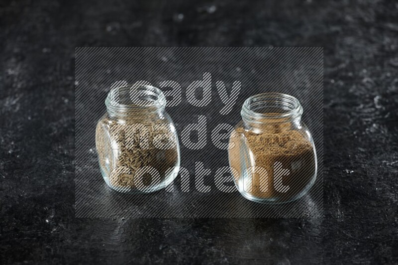 2 glass spice jars full of cumin powder and seeds on a textured black flooring